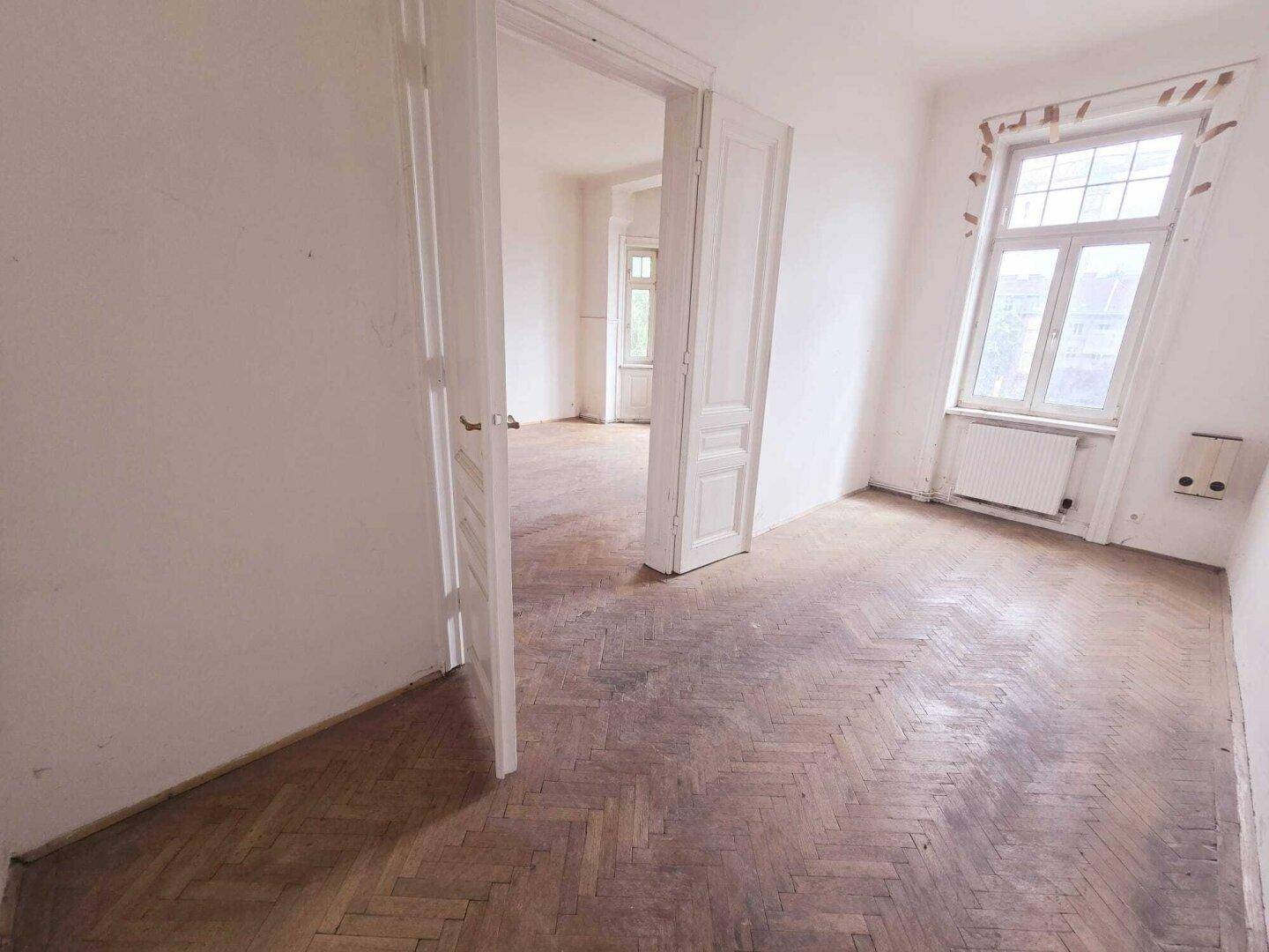 Geräumiges Zimmer mit Fischgrätparkett, hohen Decken und Zugang zu weiteren Räumen.