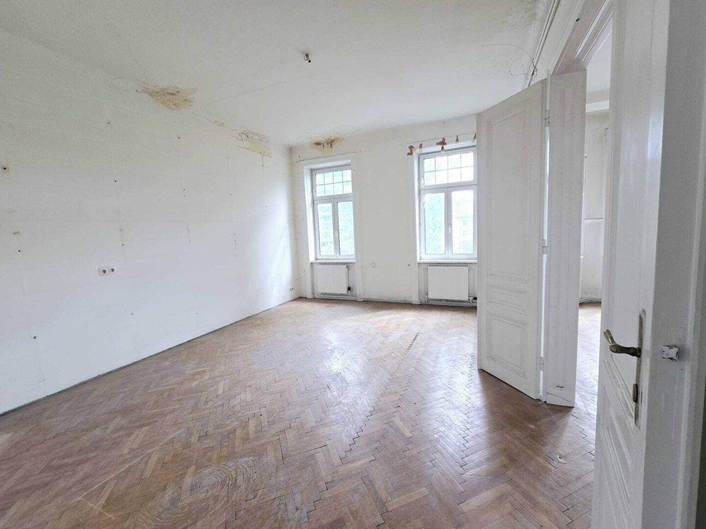 Helles Zimmer mit Fischgrätparkett und zwei Fenstern, bereit für eine Renovierung.