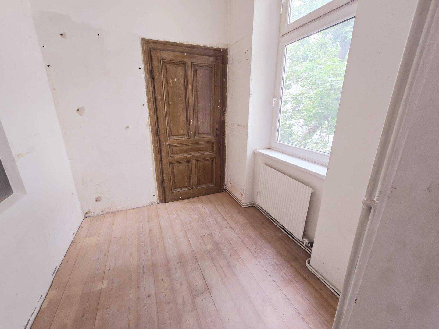 Kleines Zimmer mit hellem Dielenboden, einem Fenster und einer originalen Holztür.