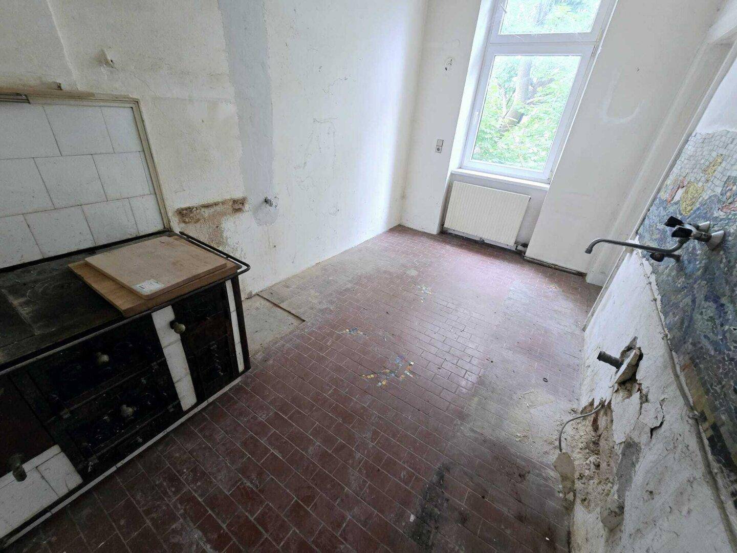 Leere Küche mit roten Fliesen, einem Fenster und einem alten Küchenherd, renovierungsbedürftig.