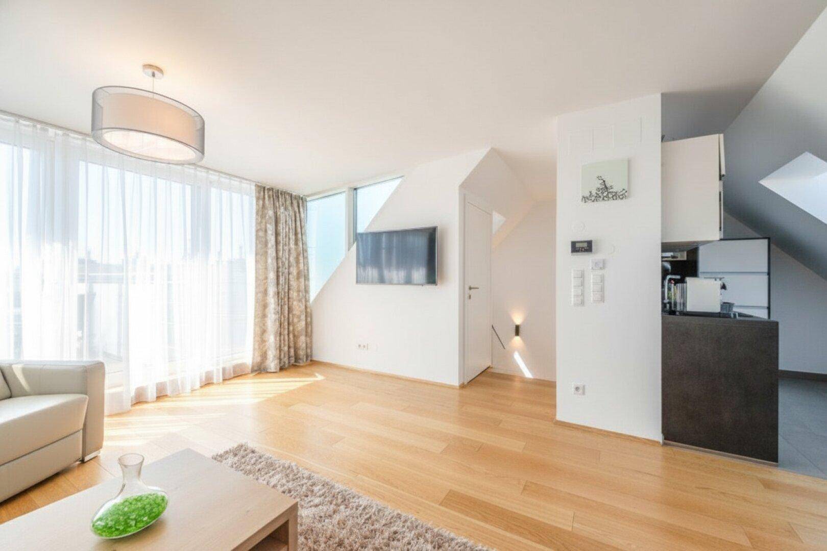 Heller Wohnbereich mit Holzboden, großen Fenstern und einem modernen TV-Gerät an der Wand.