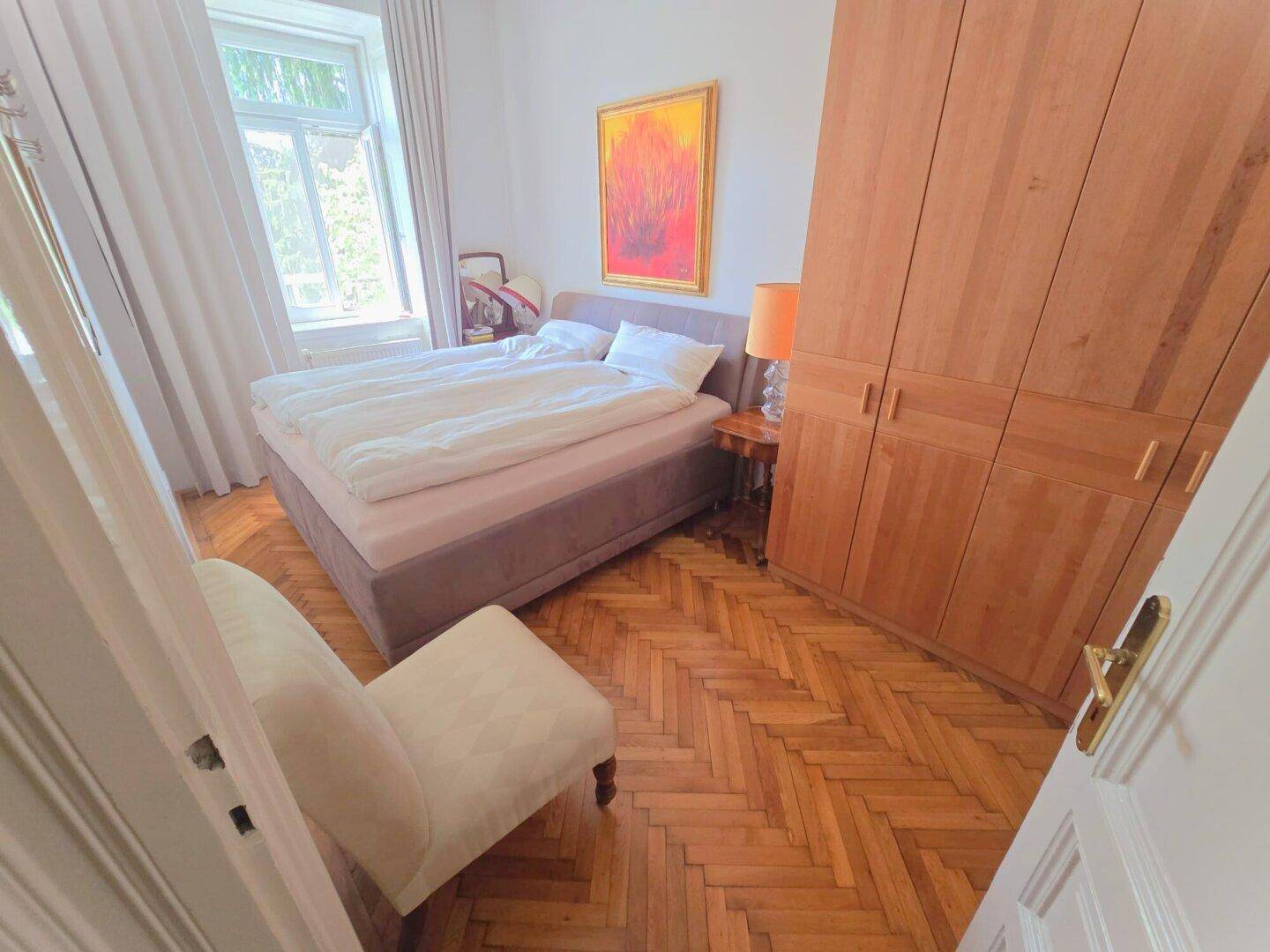 Helles Schlafzimmer mit Doppelbett, großem Holzschrank und klassischem Fischgrätparkett.