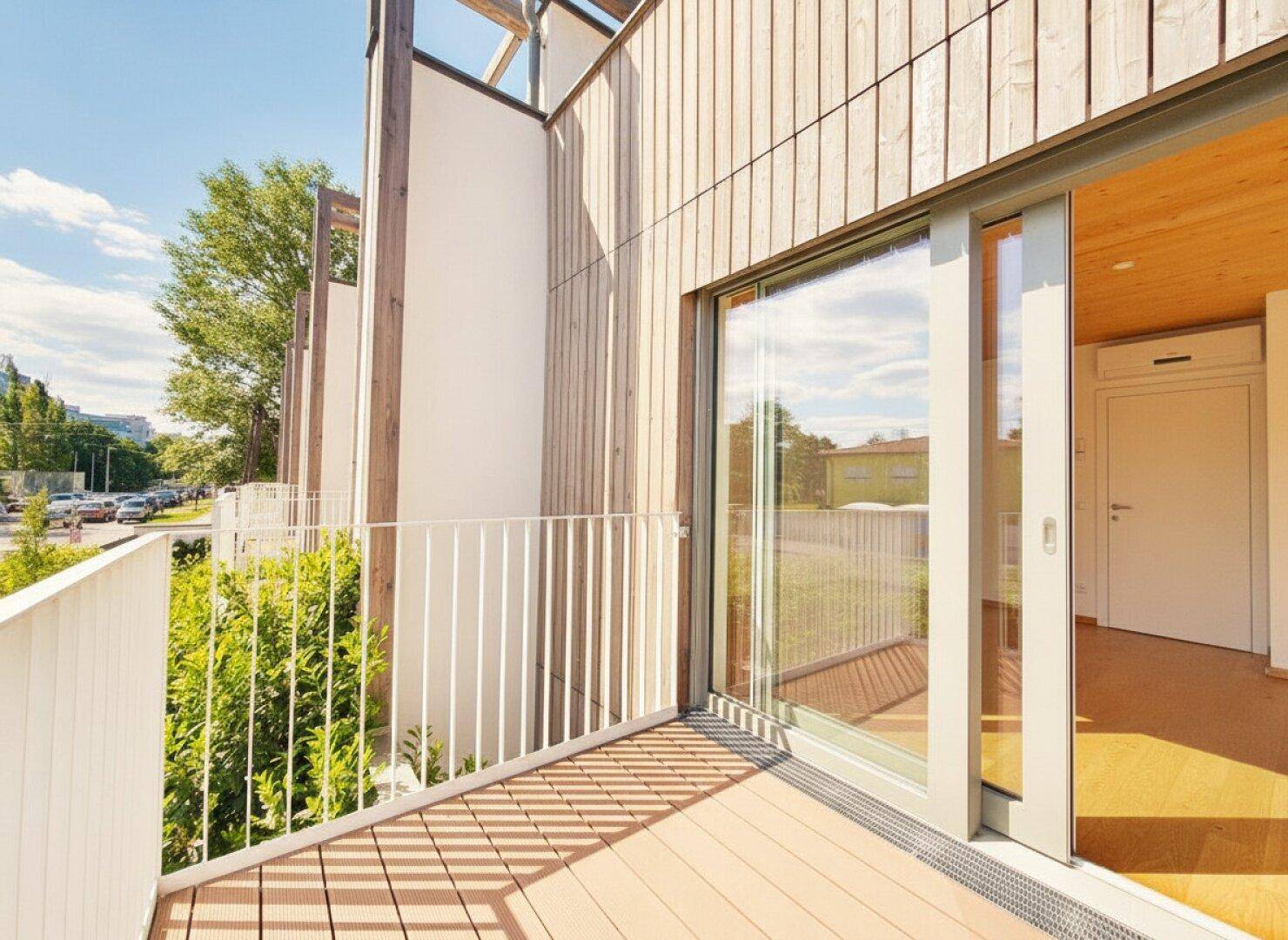 Heller Balkon mit Holzdeck, weißem Geländer und großzügiger Schiebetür zu einem modernen Gebäude.