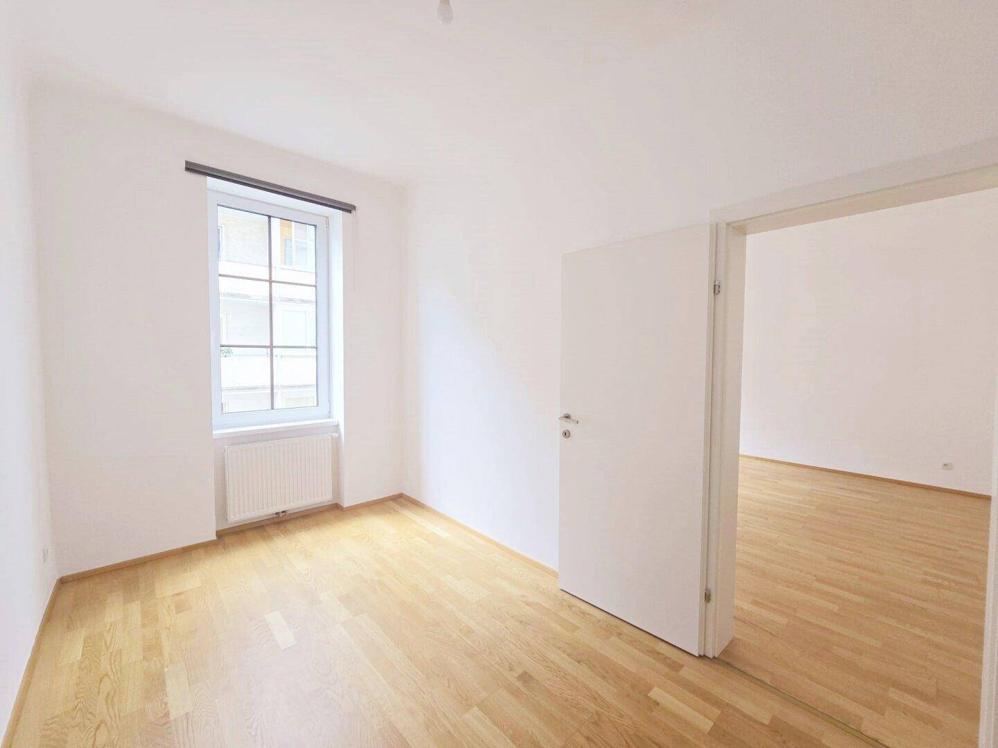 Freundliches Zimmer mit hellem Parkettboden und einem Fenster, bietet Zugang zu einem weiteren Raum.
