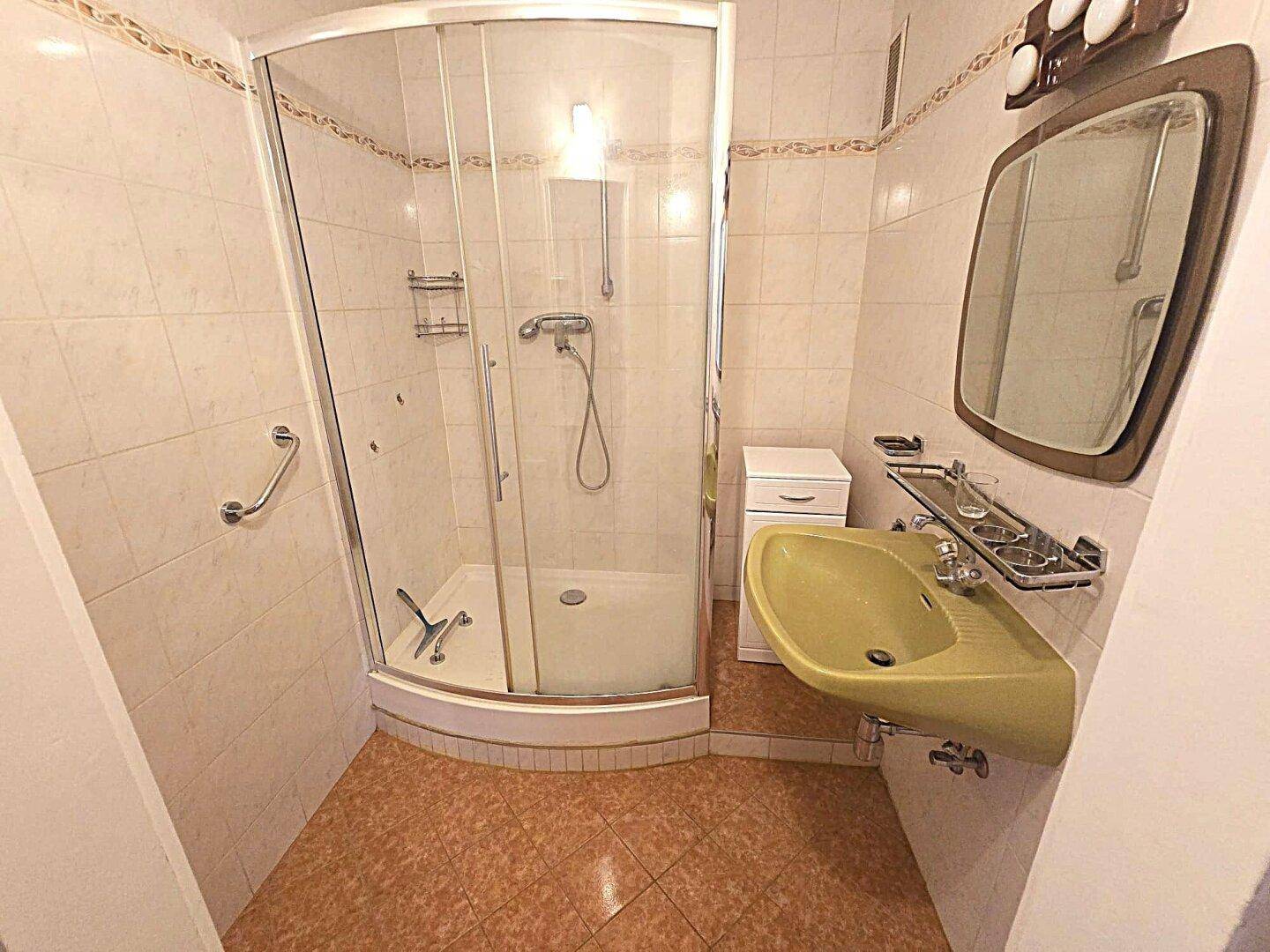 Badezimmer mit Eckdusche, gelbem Waschbecken und hellen Fliesen, ausgestattet mit Haltegriffen.