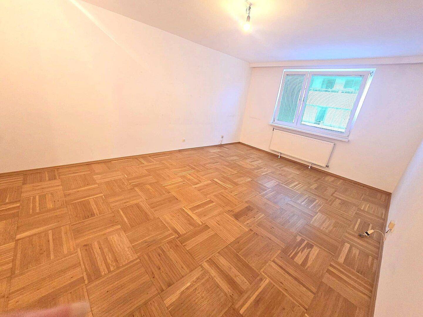 Großes, leeres Zimmer mit schönem Parkettboden und einem Fenster, das für gute Belüftung sorgt.