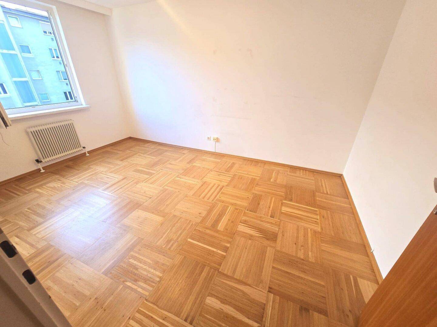Helles Zimmer mit Parkettboden und einem Fenster, das eine angenehme Atmosphäre schafft.