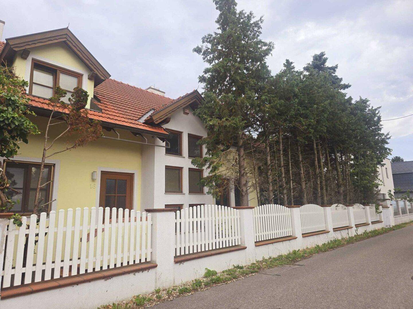 Seitenansicht des Hauses von der Straße mit weißem Lattenzaun und hohen Bäumen.