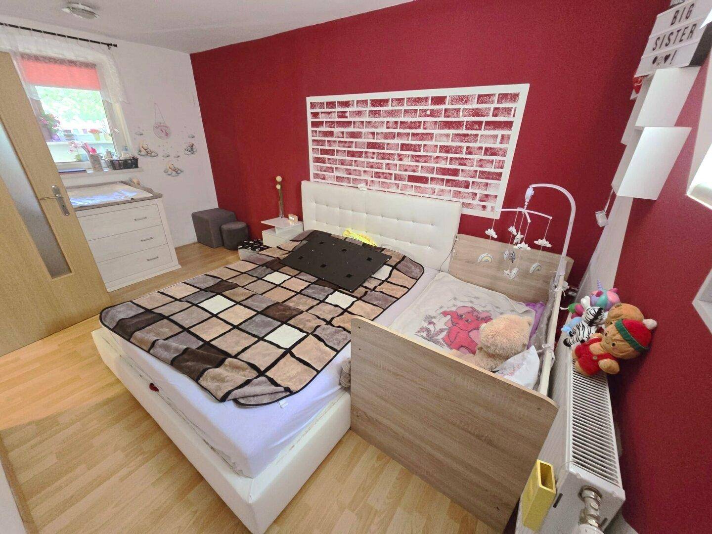 Gemütliches Schlafzimmer mit roter Akzentwand, Doppelbett und Babybett.
