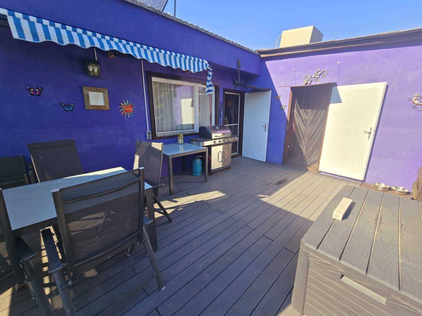Geräumige Holzterrasse mit Esstisch, Grill und einer gestreiften Markise am lila Haus.
