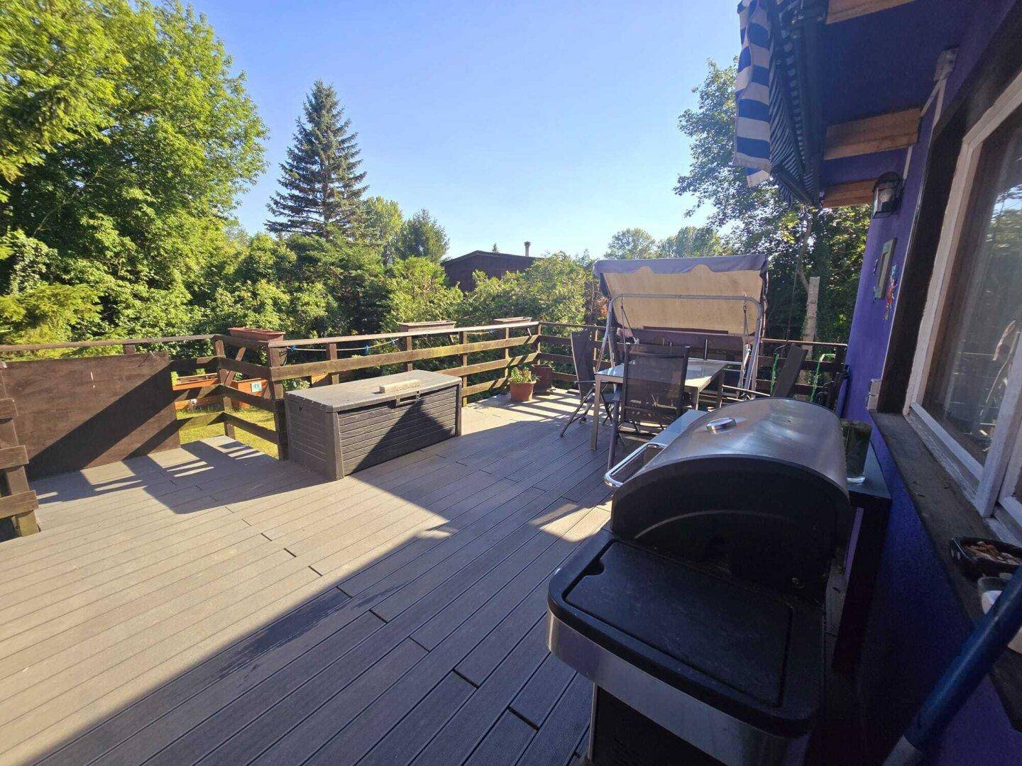 Sonnige Holzterrasse mit Grill und Stauraum, umgeben von üppigem Grün und Bäumen.