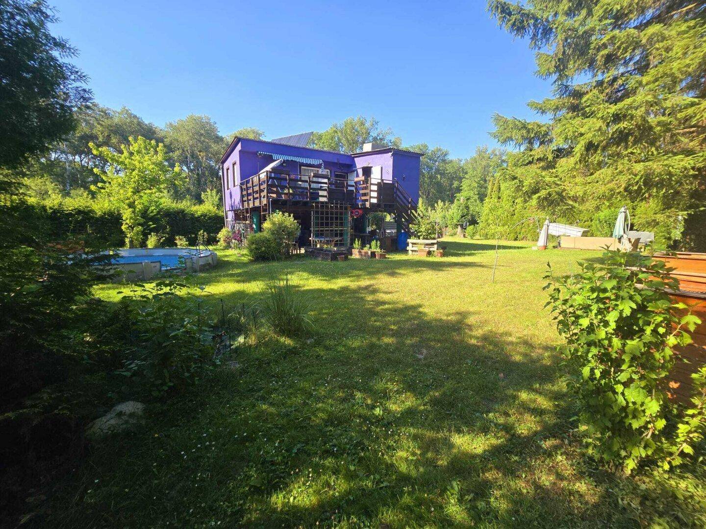 Weitläufiger Garten mit viel Grünfläche und dem markanten lila Haus unter blauem Himmel.