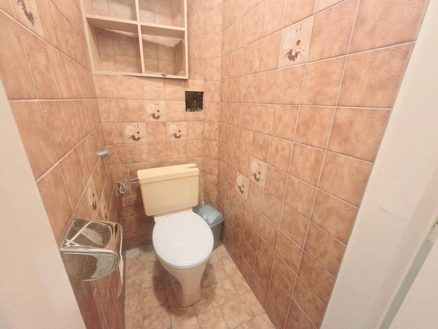 Separate Toilette mit braunen Fliesen, Fenster und einfacher Ausstattung.