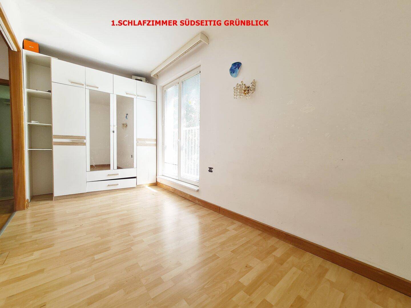 Helles Schlafzimmer mit Parkettboden, weißem Kleiderschrank und Zugang zu einem Südbalkon mit Grünblick.