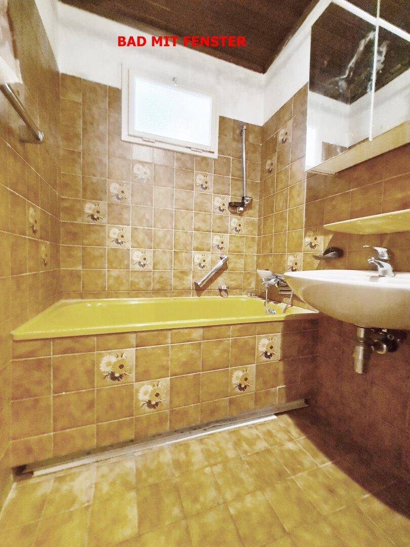 Badezimmer mit braunen Fliesen, gelber Badewanne, Waschbecken und einem Fenster für Tageslicht.