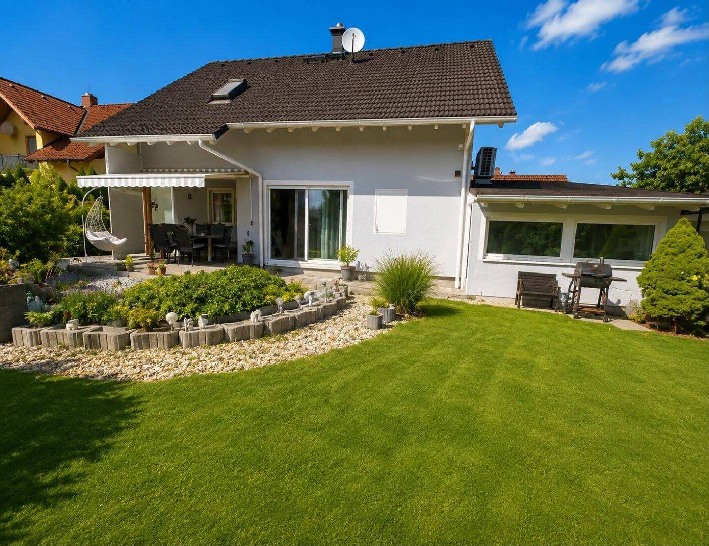 Rückansicht des Hauses mit großer Rasenfläche, Terrasse und Grillbereich unter strahlendem Sonnenschein.