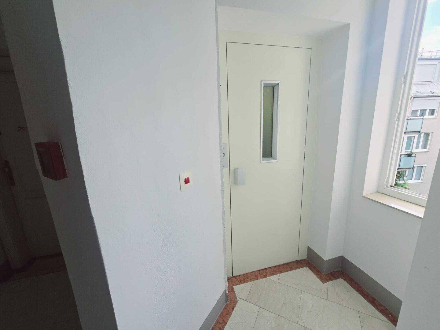Heller Flurbereich mit Aufzugstür und Fenster im Treppenhaus eines Mehrfamilienhauses.