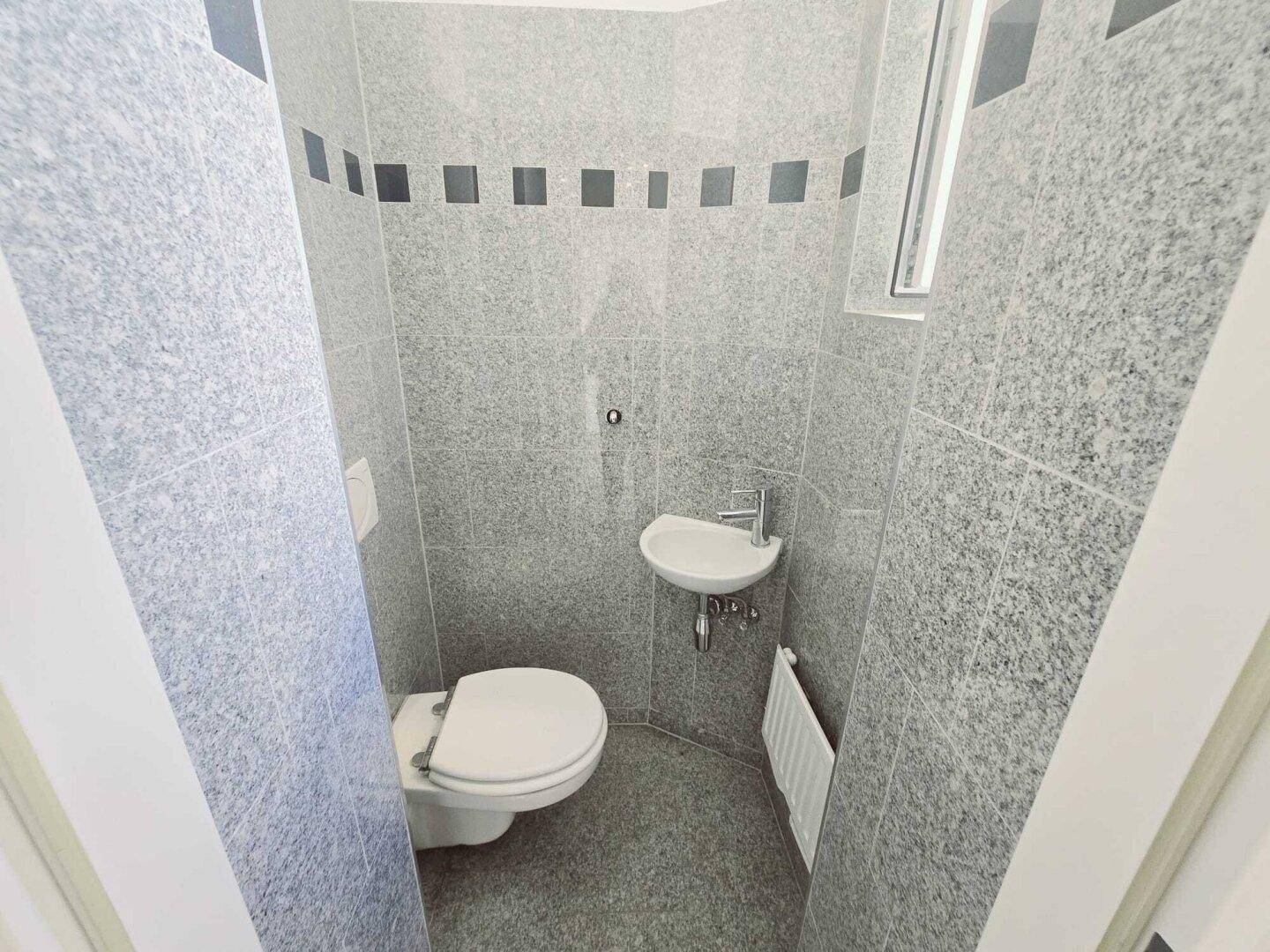 Kleines Gäste-WC mit grauen Fliesen, Toilette, Eckwaschbecken und Fenster für Tageslicht.