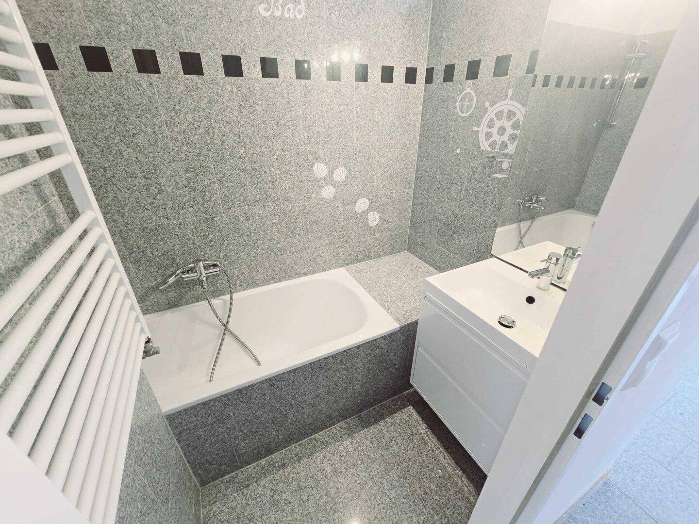 Modernes Badezimmer mit grauen Fliesen, Badewanne, Einzelwaschbecken und Handtuchheizkörper.