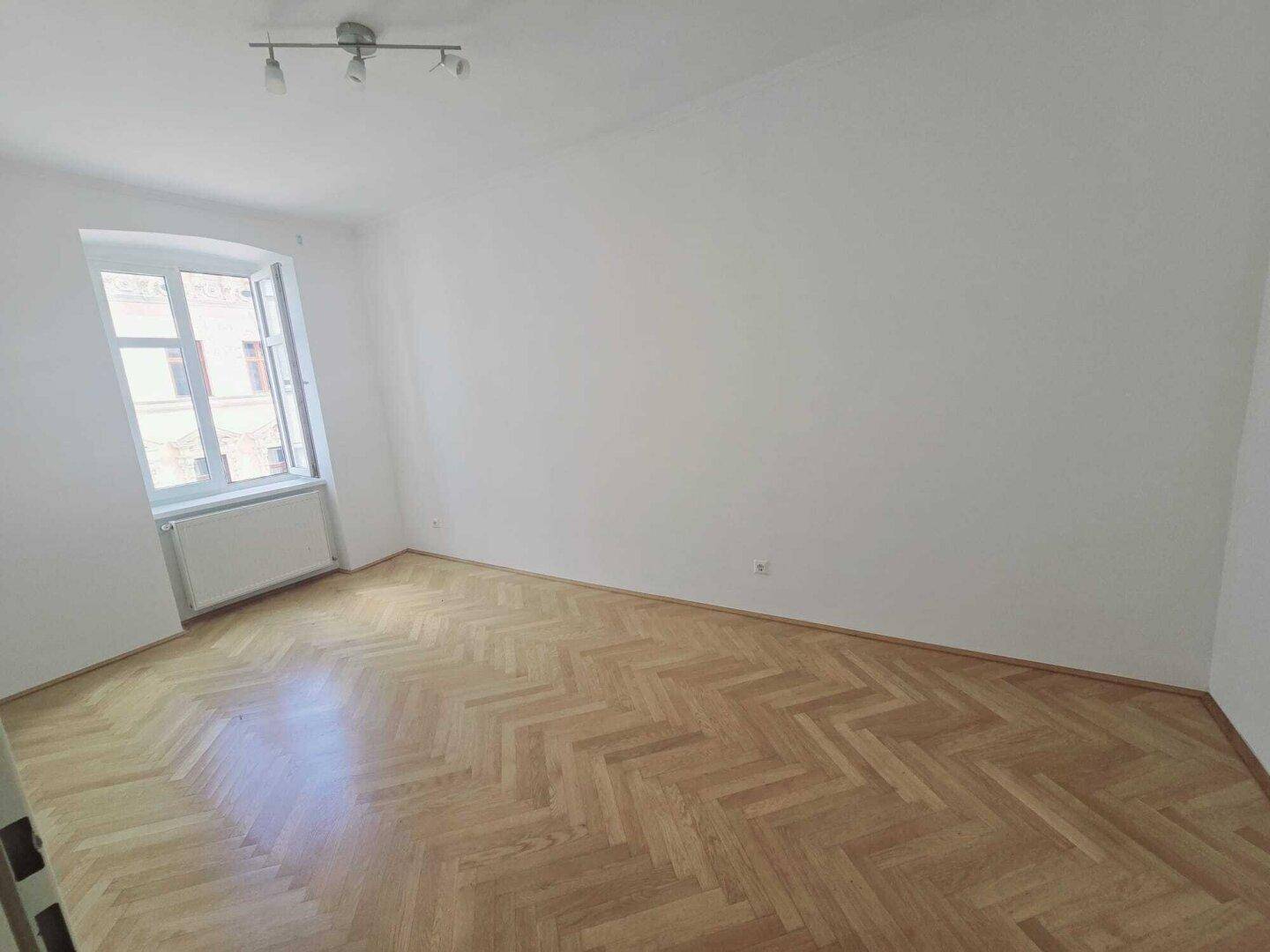 Leeres Zimmer mit Parkettboden und einem großen Fenster, das viel Tageslicht hereinlässt.