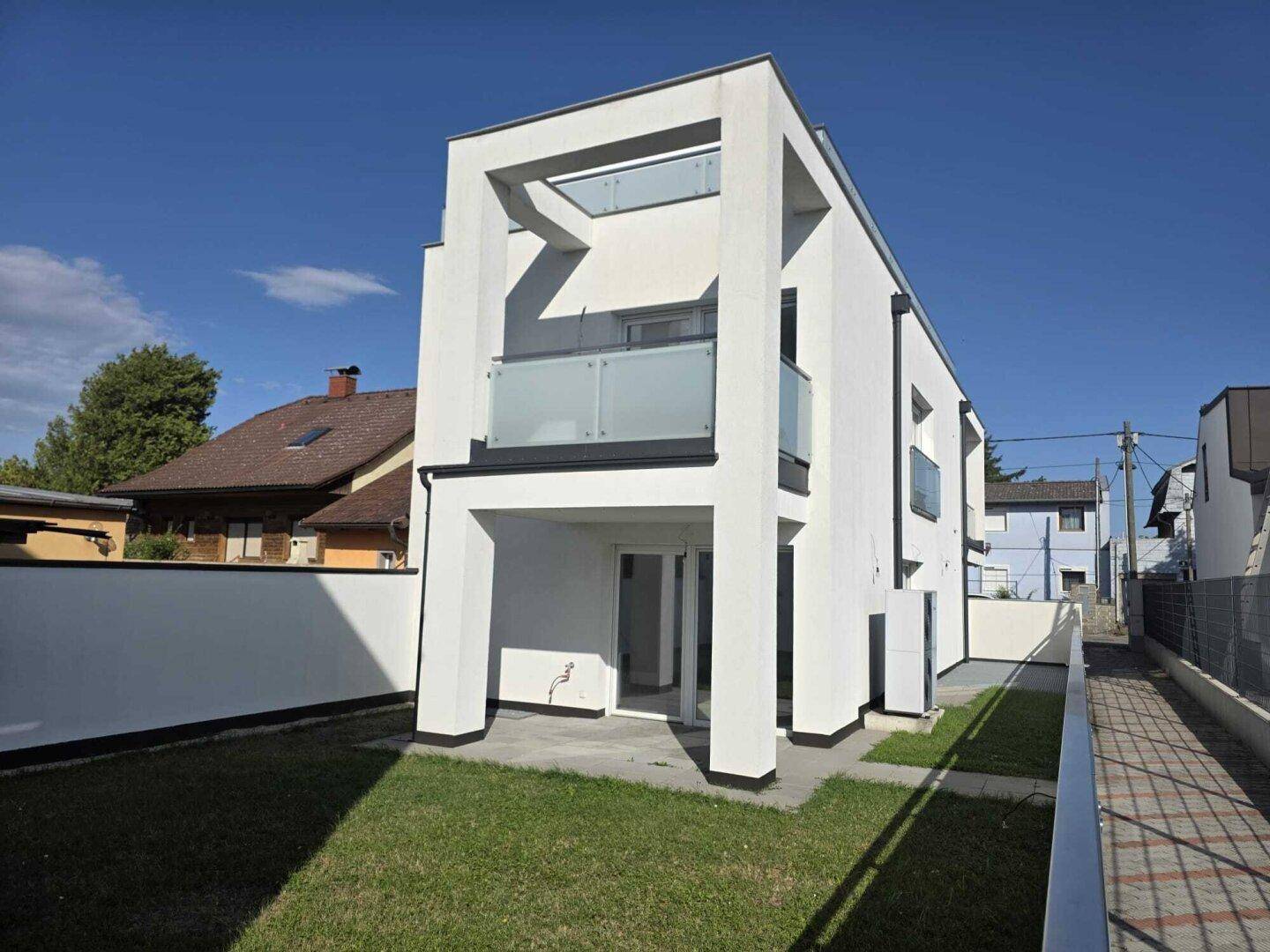 Neubau mit weißer Fassade, Balkonen und einem kleinen Rasengarten unter strahlendem Himmel.