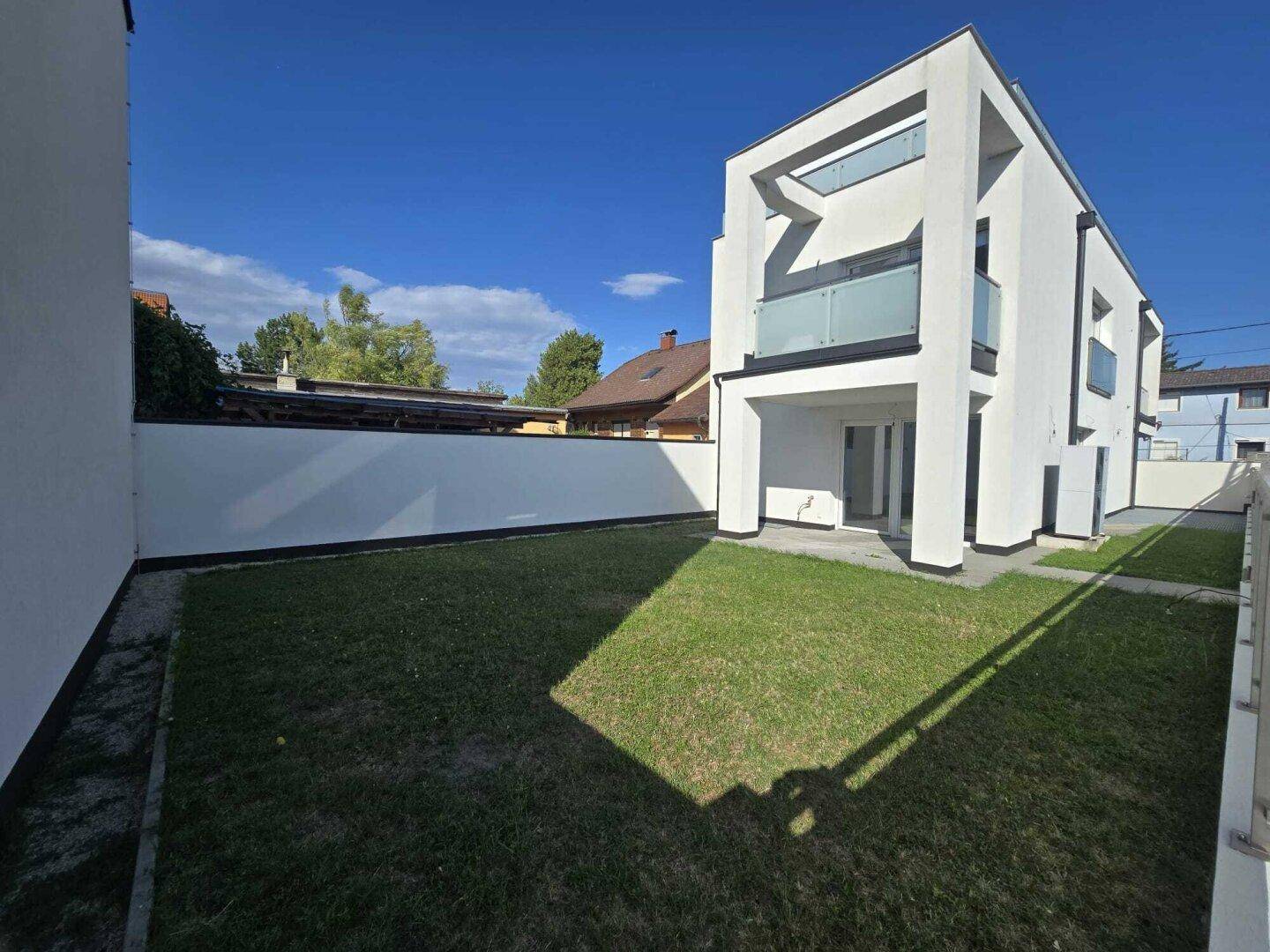 Seitlicher Gartenbereich mit Rasenfläche und moderner Gebäudefassade unter blauem Himmel.