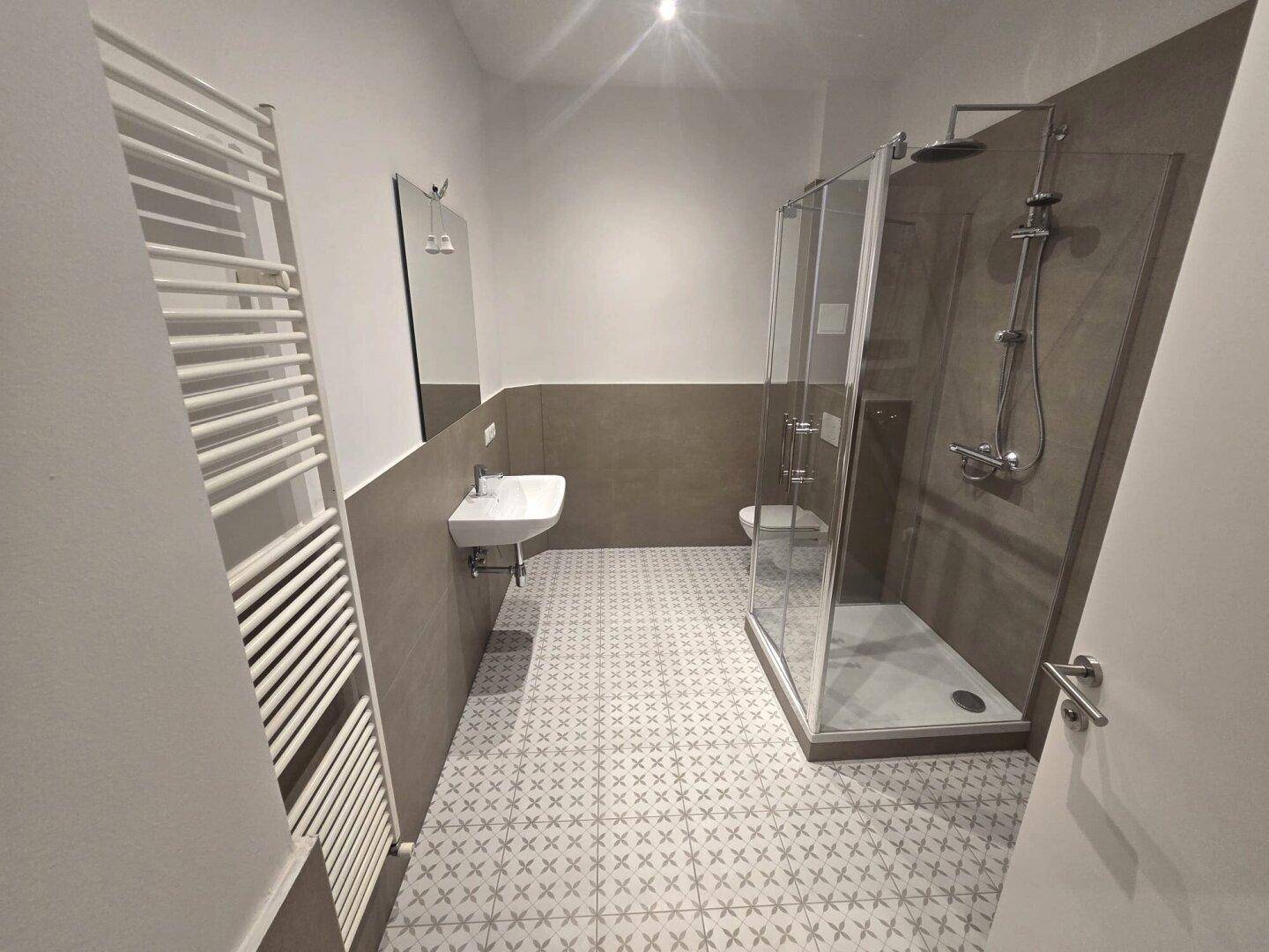 Modernes Badezimmer mit begehbarer Dusche, Wand-WC, Waschbecken und Handtuchheizkörper, in Grautönen gefliest.