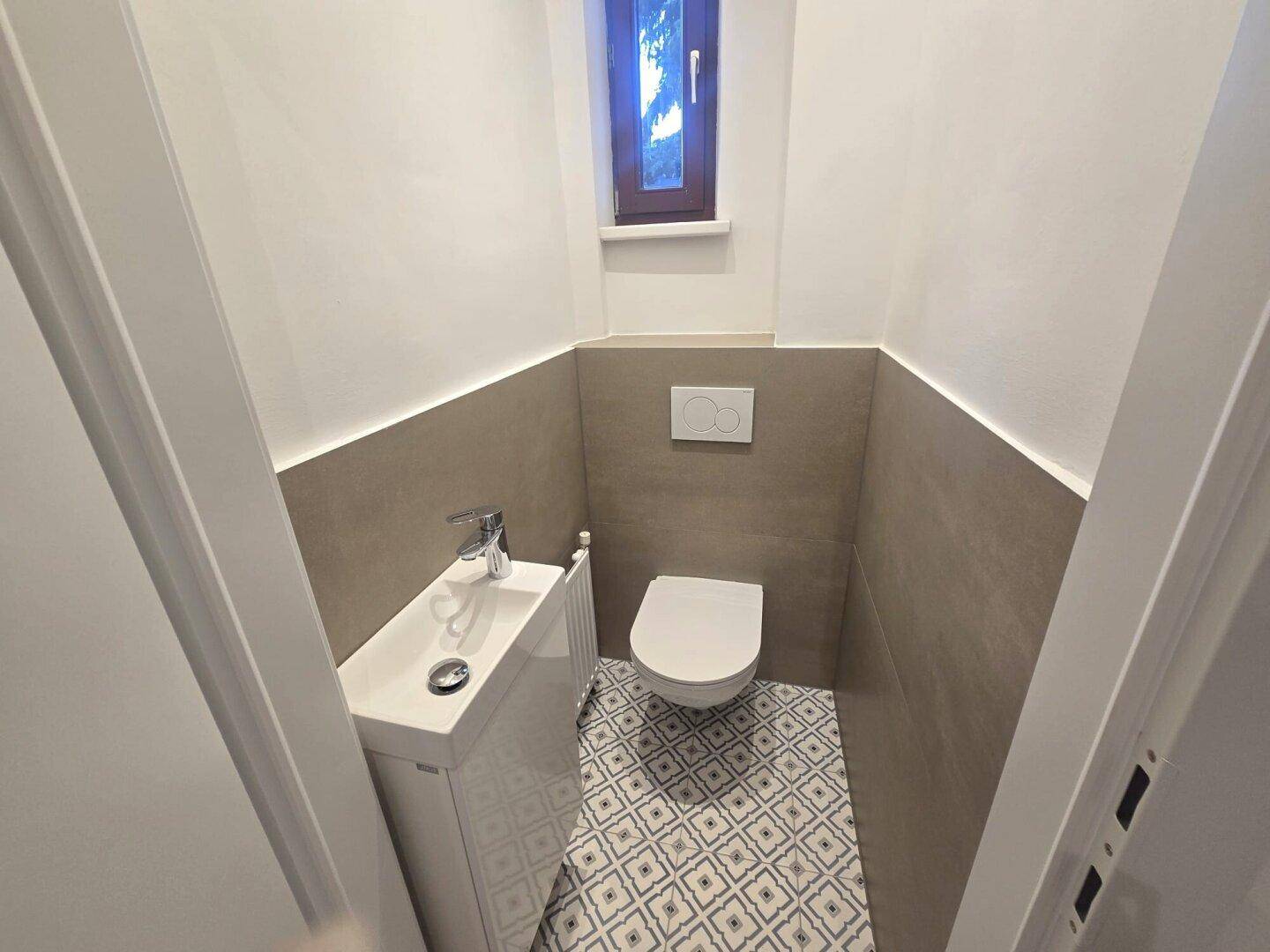 Kompaktes Gäste-WC mit Wand-WC, kleinem Waschbecken und Fenster, modern gefliest.