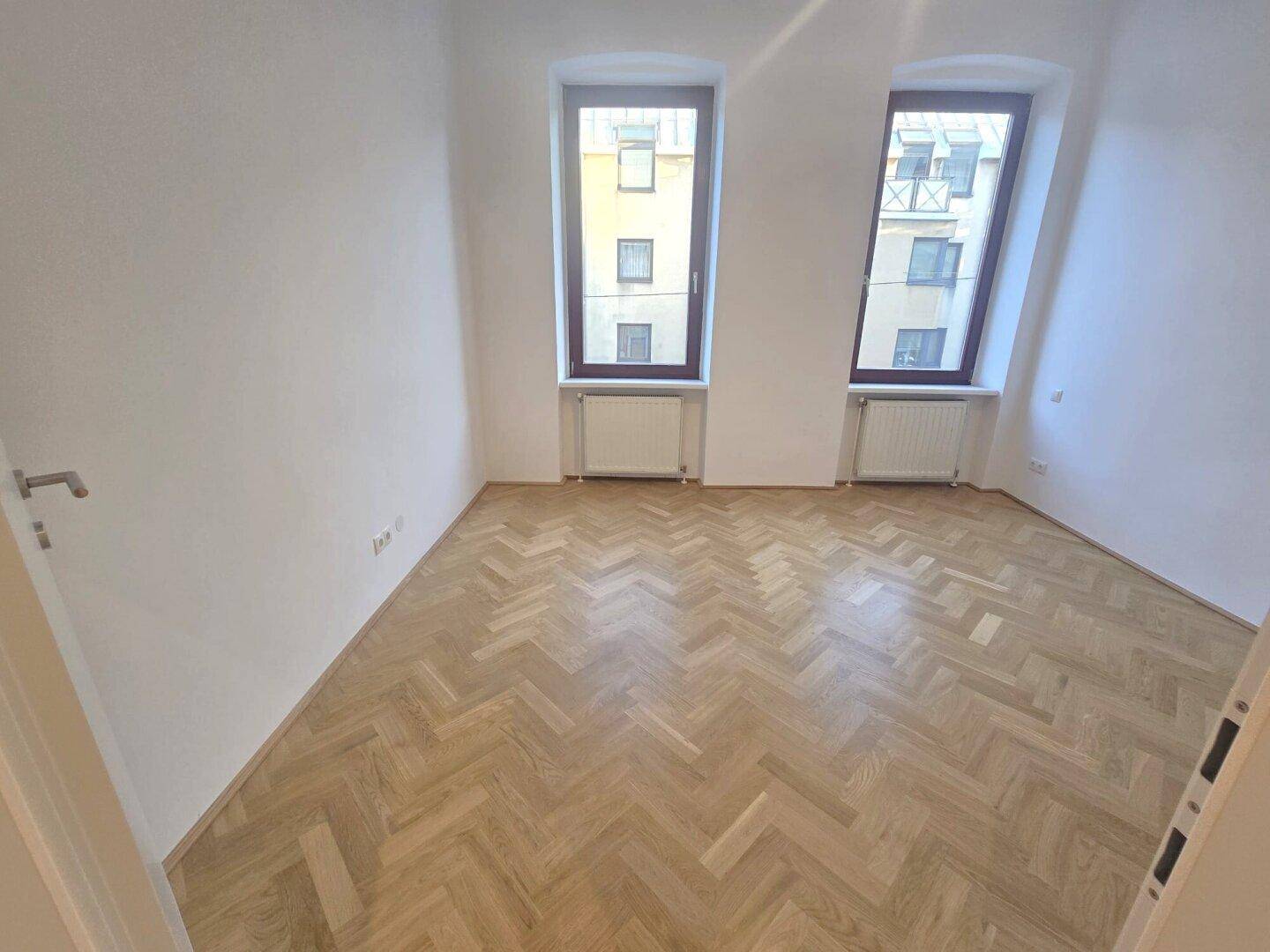 Geräumiges Zimmer mit Fischgrätparkett und zwei Fenstern, die viel Tageslicht hereinlassen.