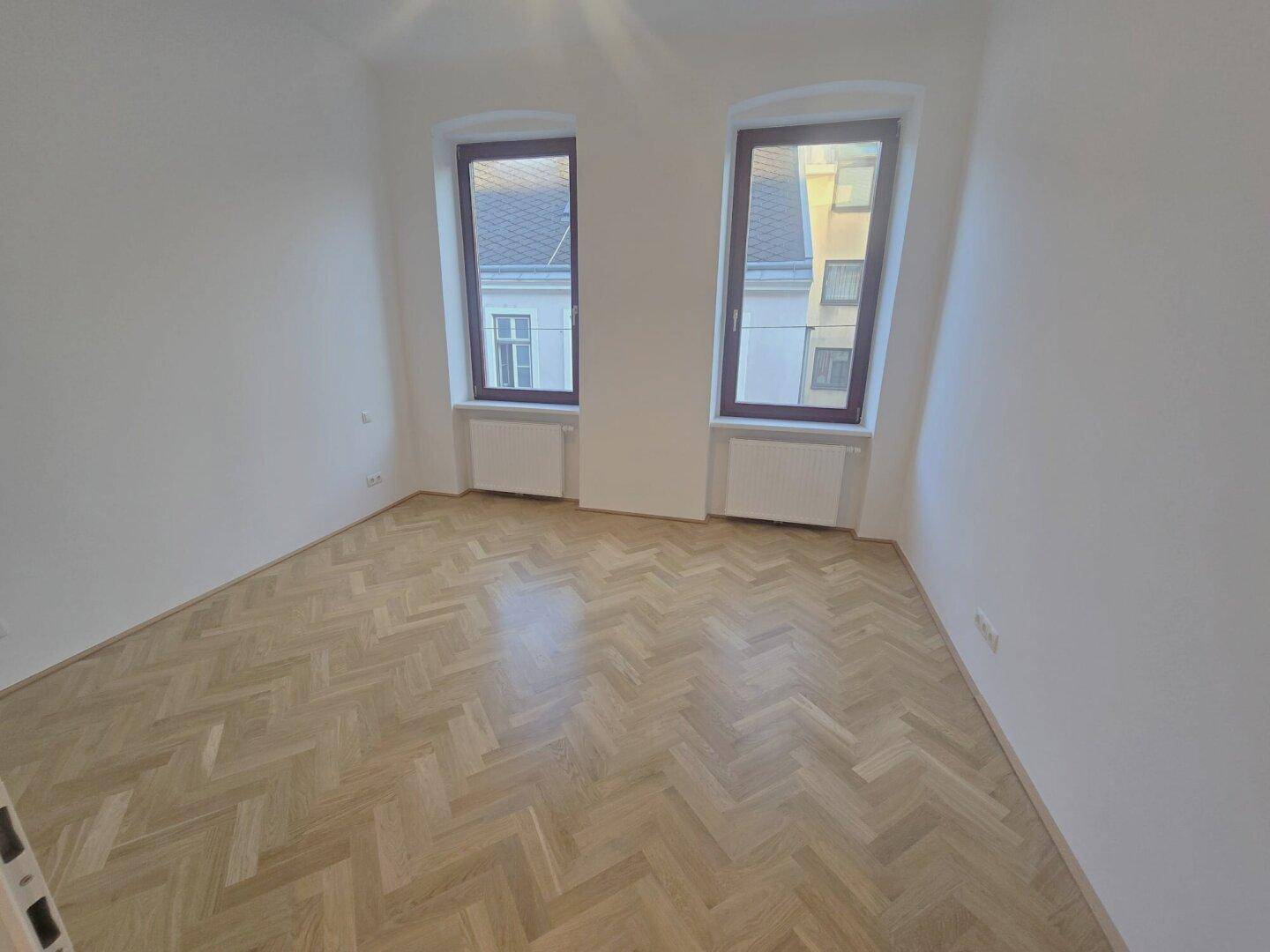 Helles Zimmer mit Fischgrätparkett und zwei Fenstern, die einen Blick auf die Umgebung bieten.