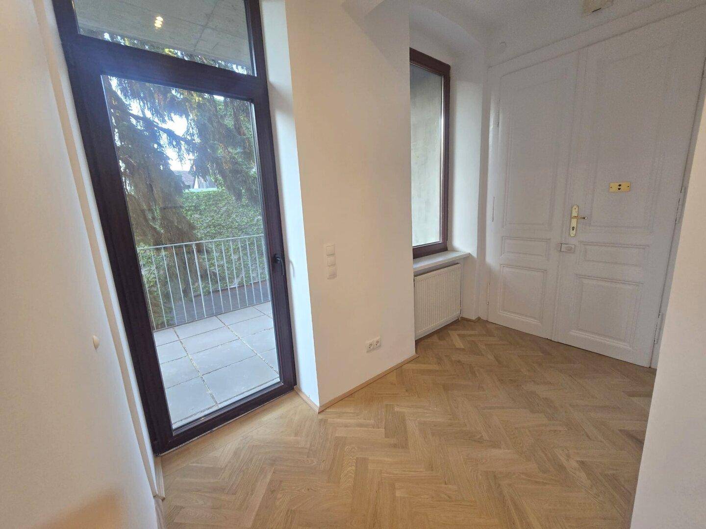 Wohnraum mit Fischgrätparkett und großer Glastür, die direkten Zugang zur Außenterrasse bietet.