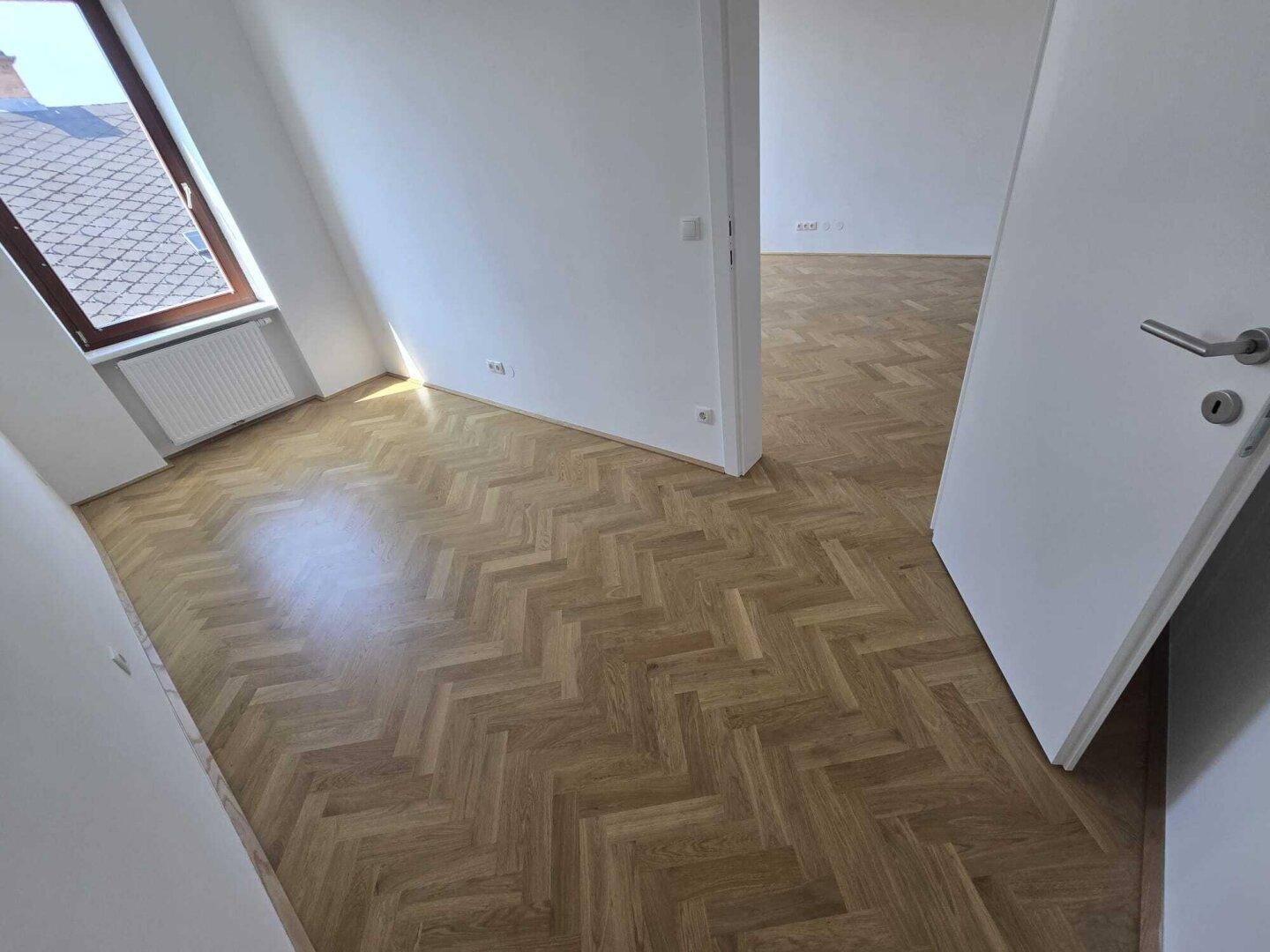 Helles Zimmer mit Fischgrätparkett, weißen Wänden und einem Fenster, das den Raum erhellt.