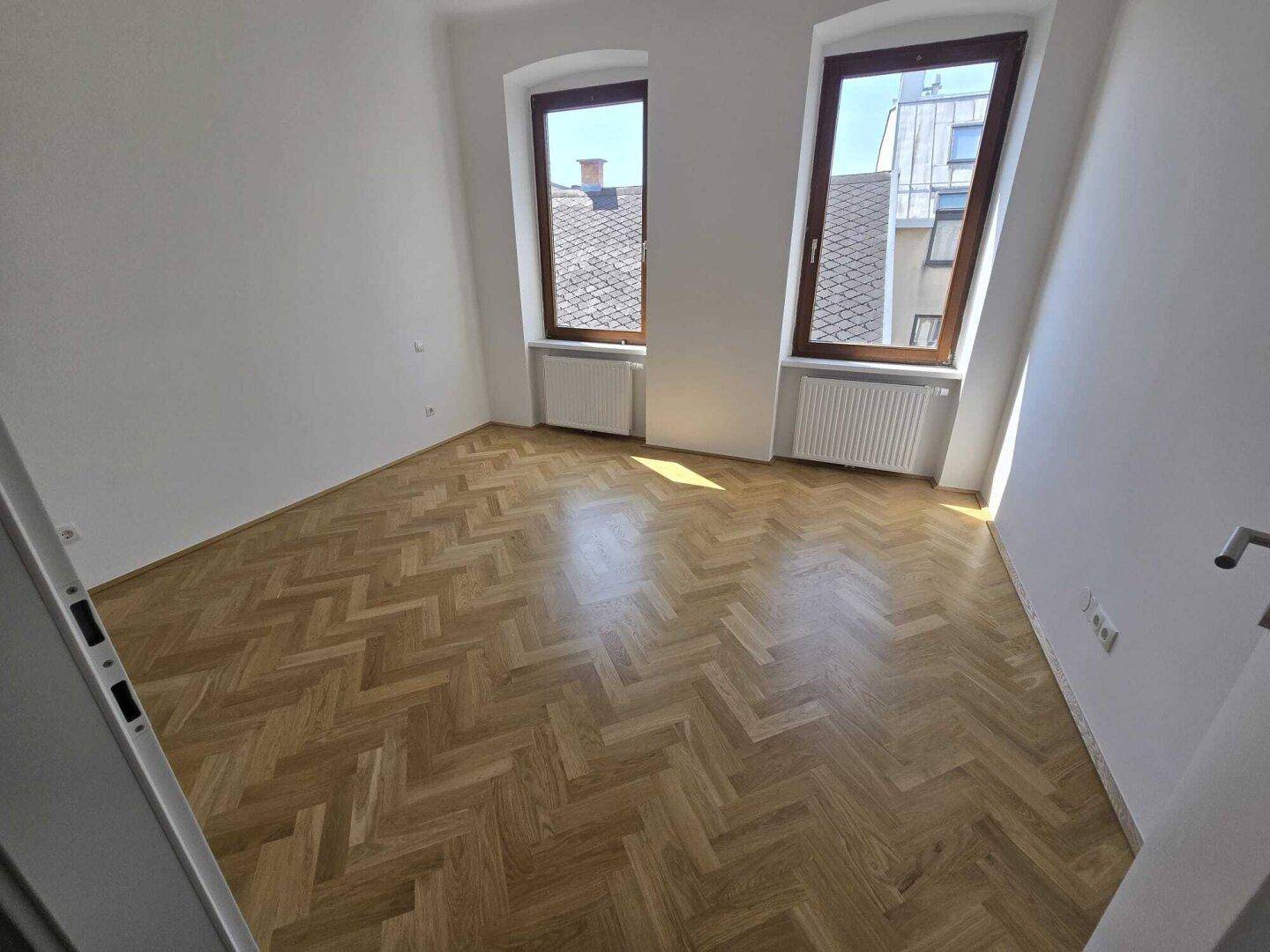 Helles Schlafzimmer mit Fischgrätparkett, weißen Wänden und zwei Fenstern mit Blick auf die Dächer.