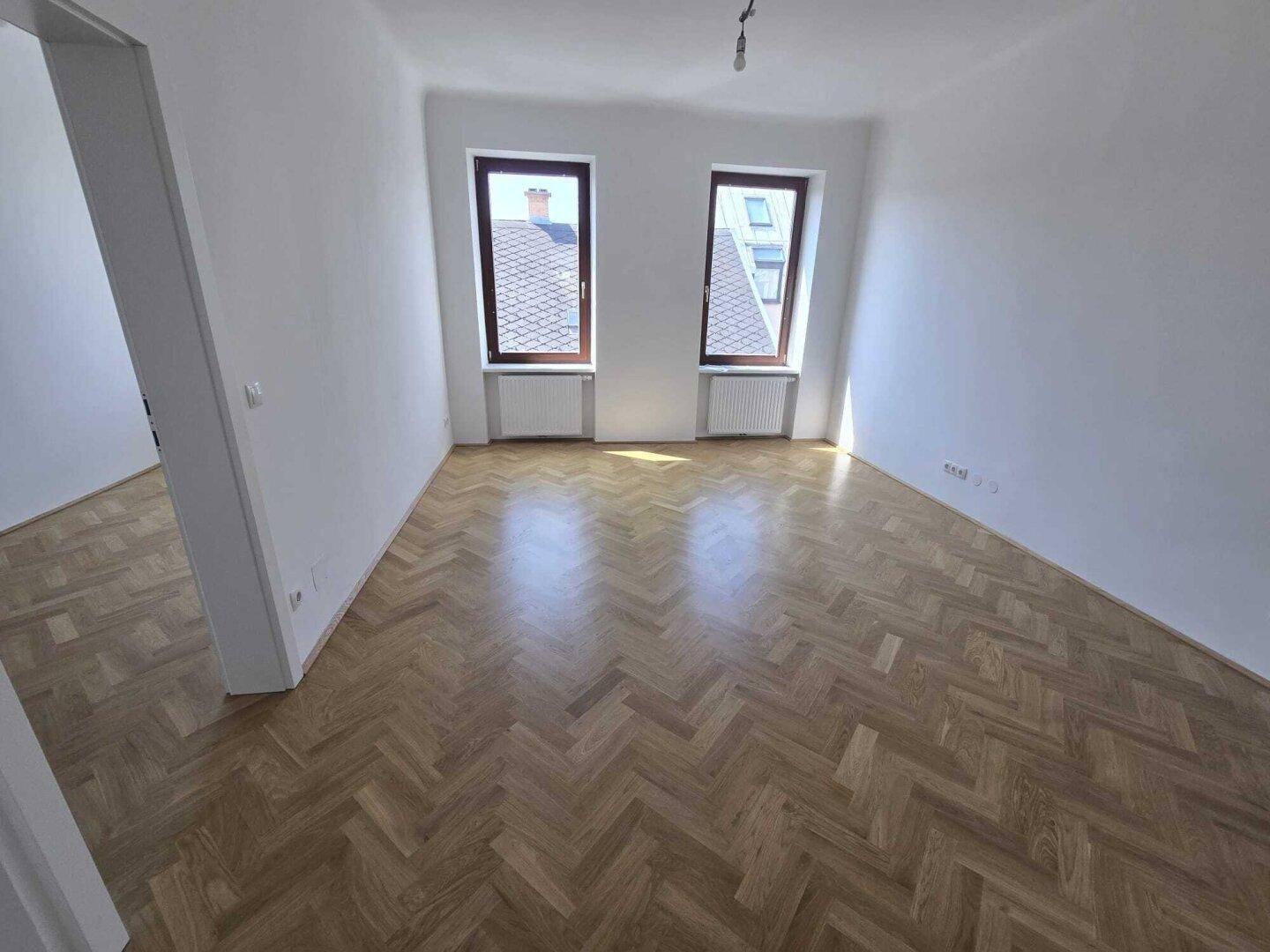 Geräumiges Wohnzimmer mit Fischgrätparkett, weißen Wänden und zwei Fenstern, die viel Licht hereinlassen.
