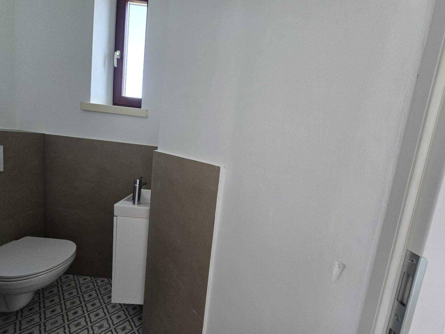 Kleines Gäste-WC mit Toilette, wandhängendem Waschbecken und einem schmalen Fenster für Tageslicht.