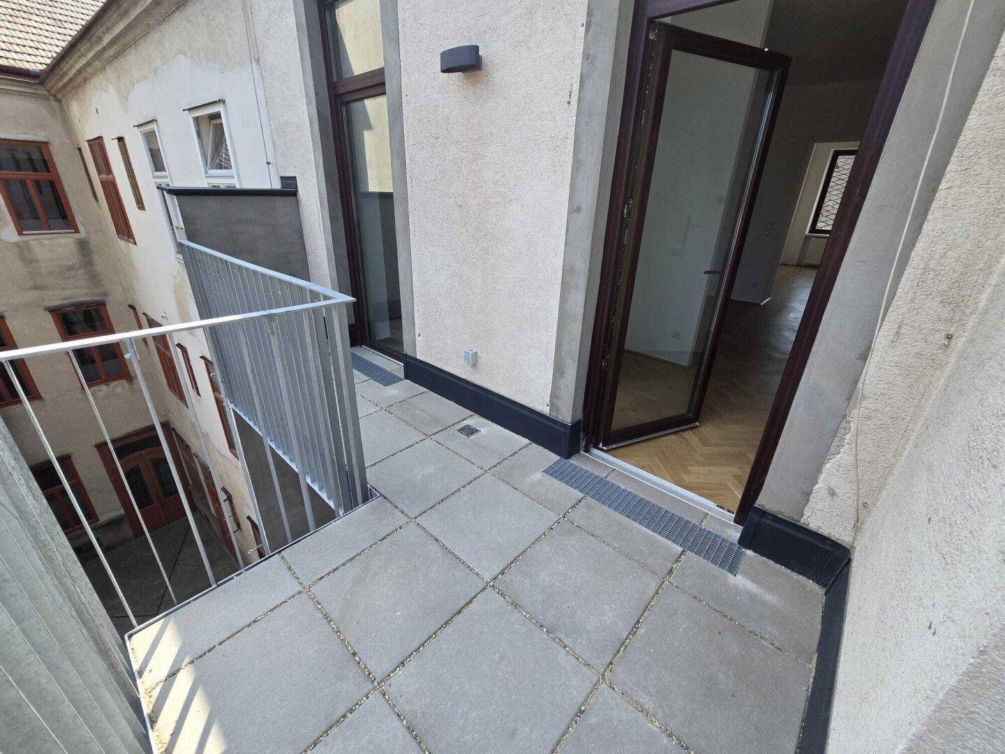 Balkon mit grauen Fliesen und Metallgeländer, mit Blick auf einen Innenhof und Zugang durch eine Glastür.