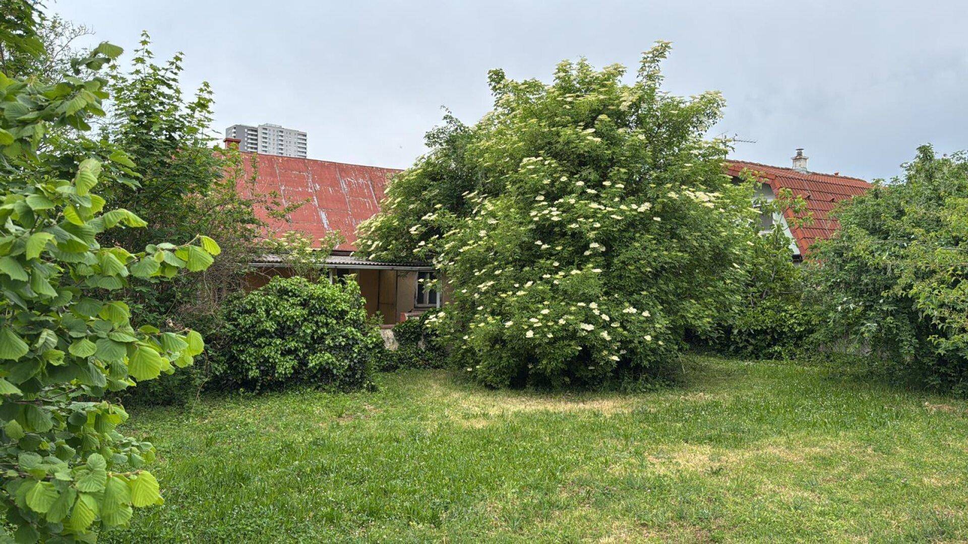Üppiger Garten mit blühenden Bäumen und Sträuchern, der das Haus teilweise verdeckt, unter bewölktem Himmel.