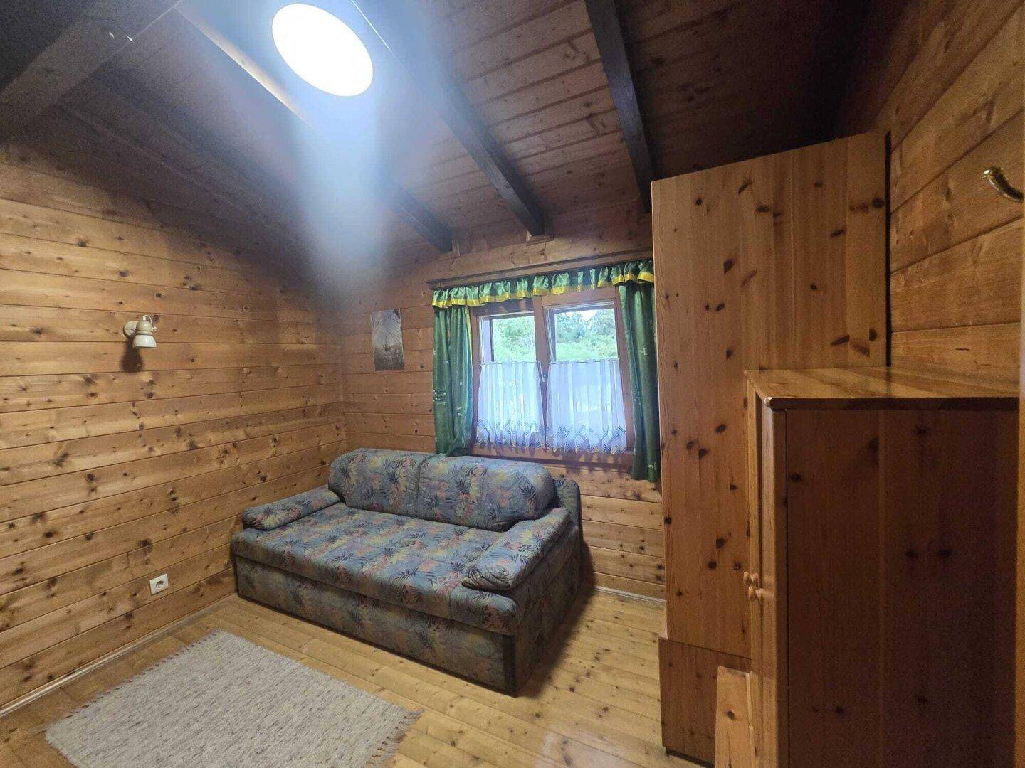 Schlafzimmer mit Holzwänden, Holzbalkendecke, Schlafsofa und einem Fenster mit Vorhängen.
