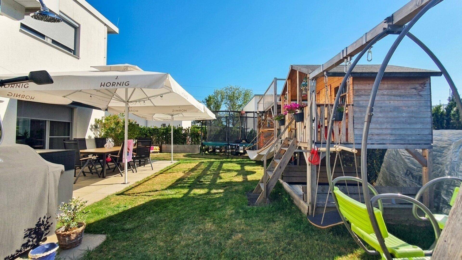 Großer Garten mit Terrasse, Spielplatz, Trampolin und Sitzgelegenheiten unter Sonnenschirm.