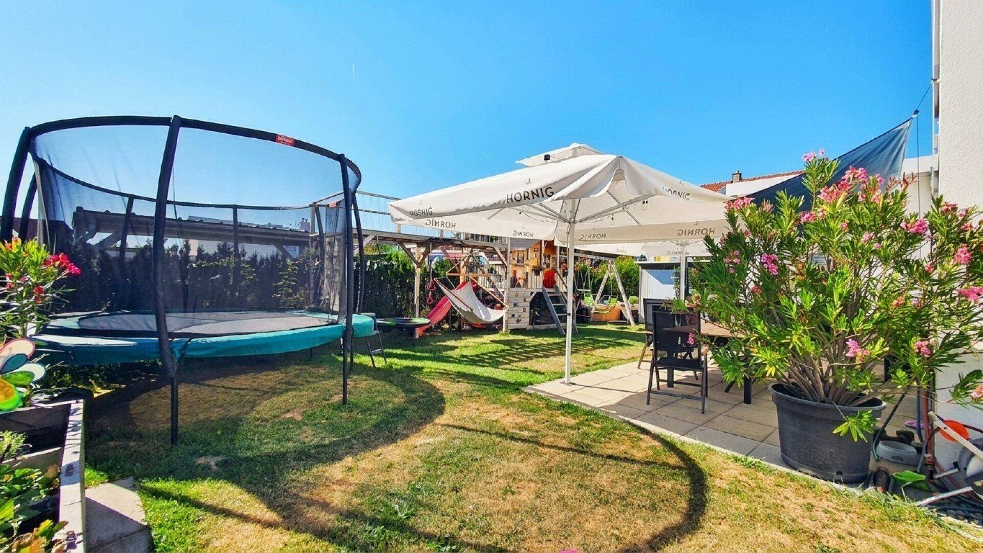 Sonniger Gartenbereich mit großem Trampolin, Terrasse und blühenden Pflanzen.