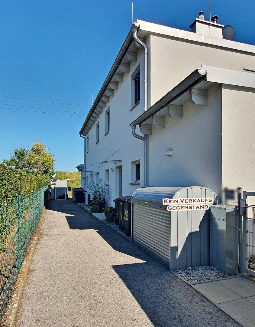 Seitenansicht eines Wohnhauses mit gepflastertem Weg und Mülltonnenbox.