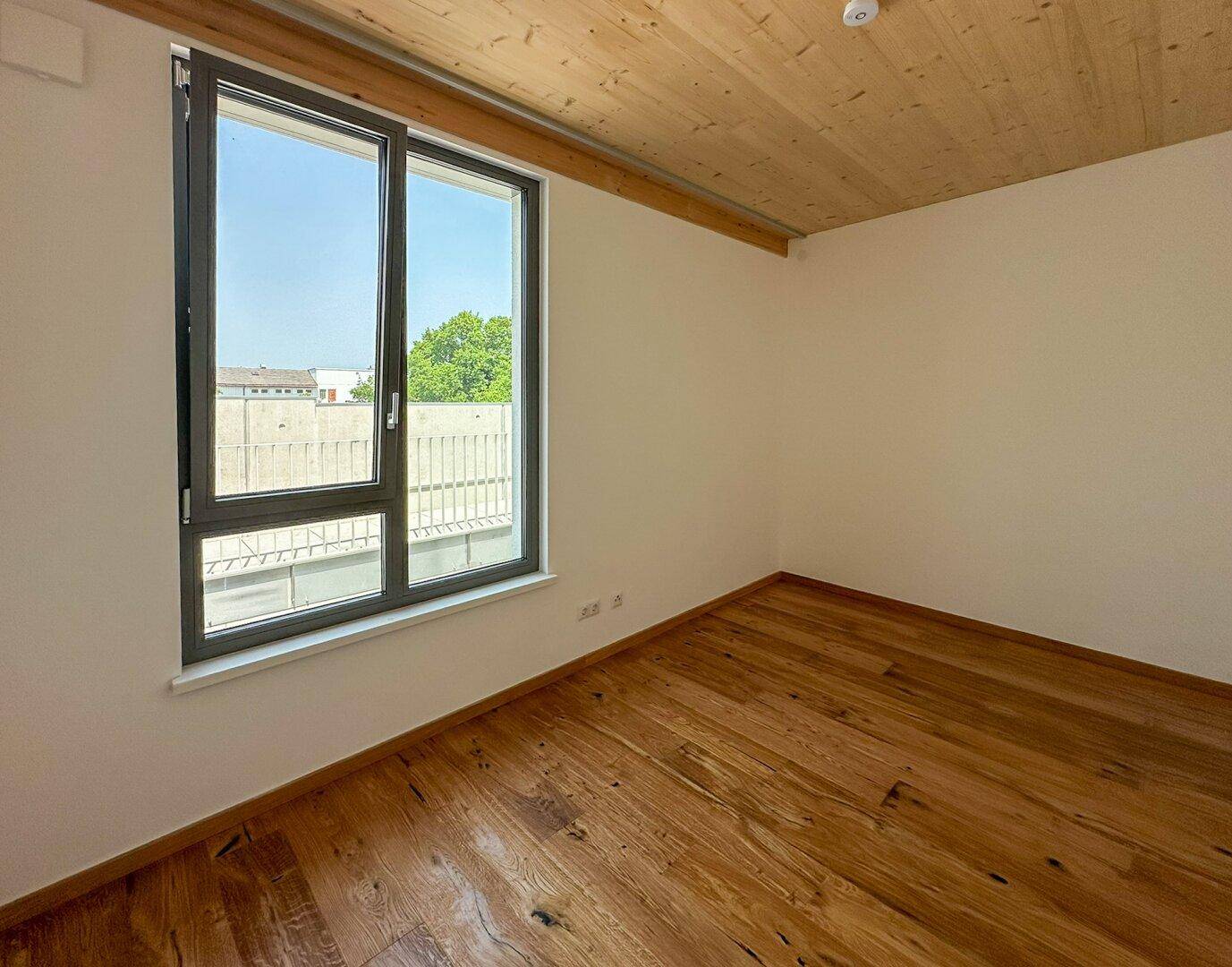 Helles Schlafzimmer mit Holzboden, Holzdecke und großem Fenster mit Blick ins Grüne.