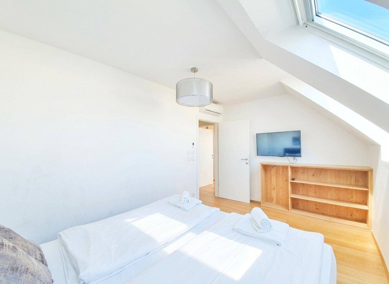 Helles Schlafzimmer mit Doppelbett, Holzboden, Dachfenster und einem an der Wand montierten Fernseher.