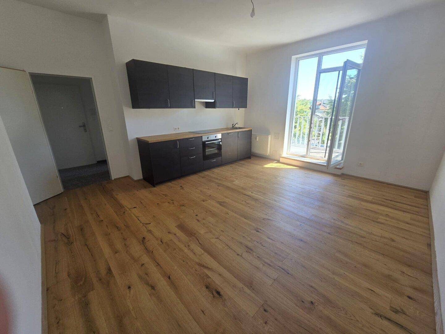 Heller Wohnbereich mit Holzboden und integrierter Küchenzeile, mit Zugang zu einem Balkon.