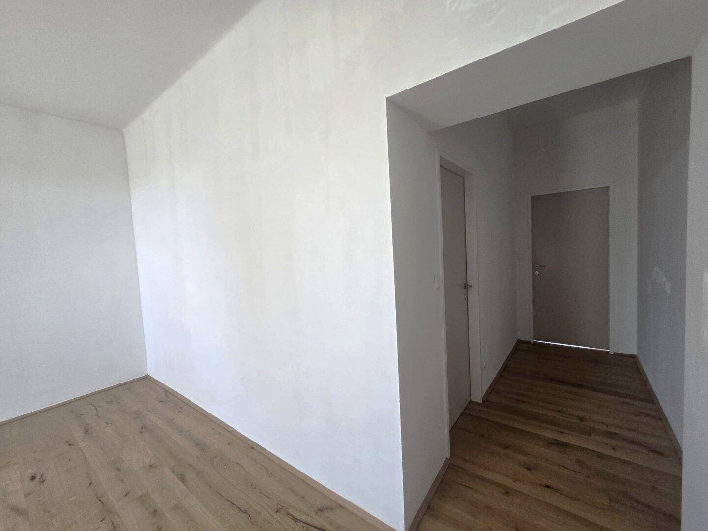Leerer Raum mit Holzboden und weiß gestrichenen Wänden, mit Zugang zu einem Flur.