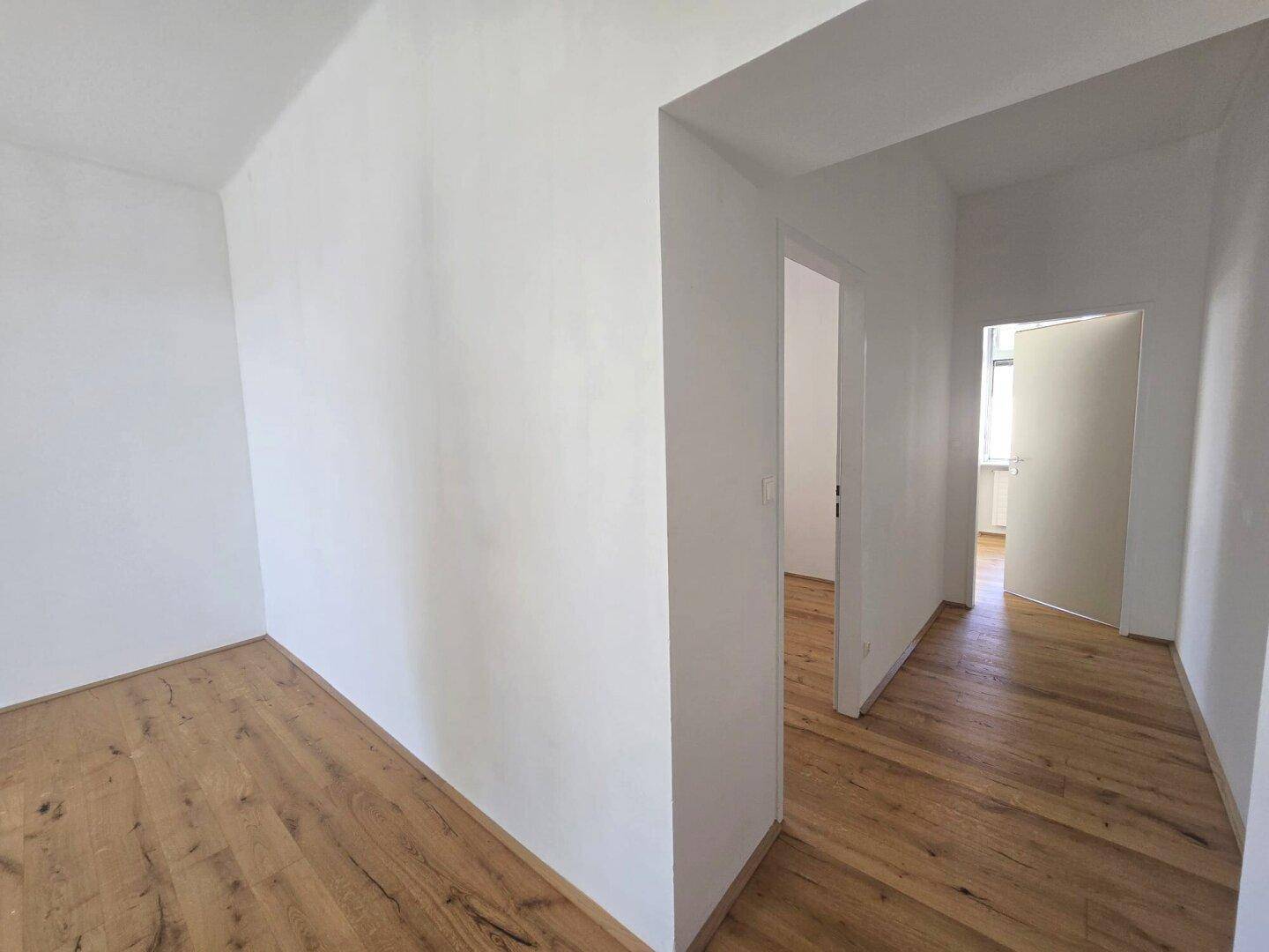 Leerer Raum mit Holzboden und weiß gestrichenen Wänden, führt in einen Flur.