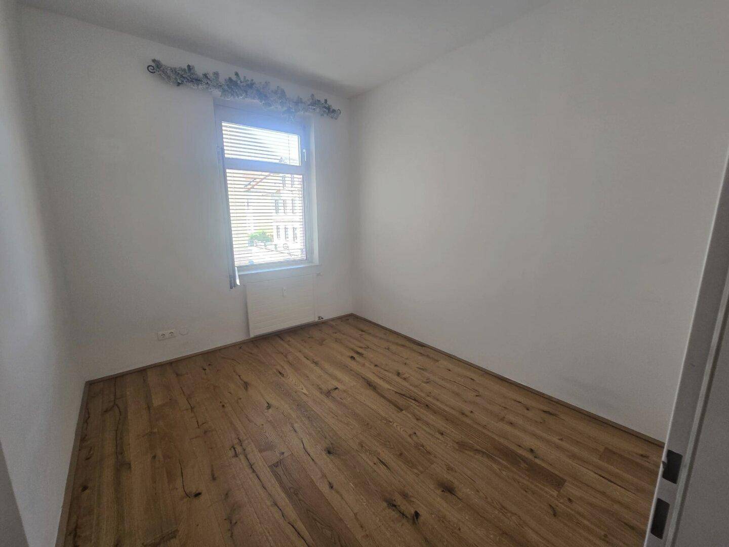 Kleines, helles Zimmer mit Holzboden und einem Fenster, ideal als Schlaf- oder Arbeitszimmer.