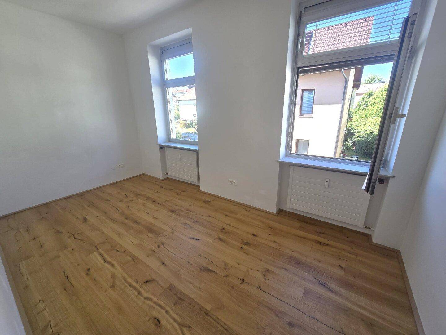 Helles Zimmer mit Holzboden und zwei Fenstern, die viel Tageslicht hereinlassen.