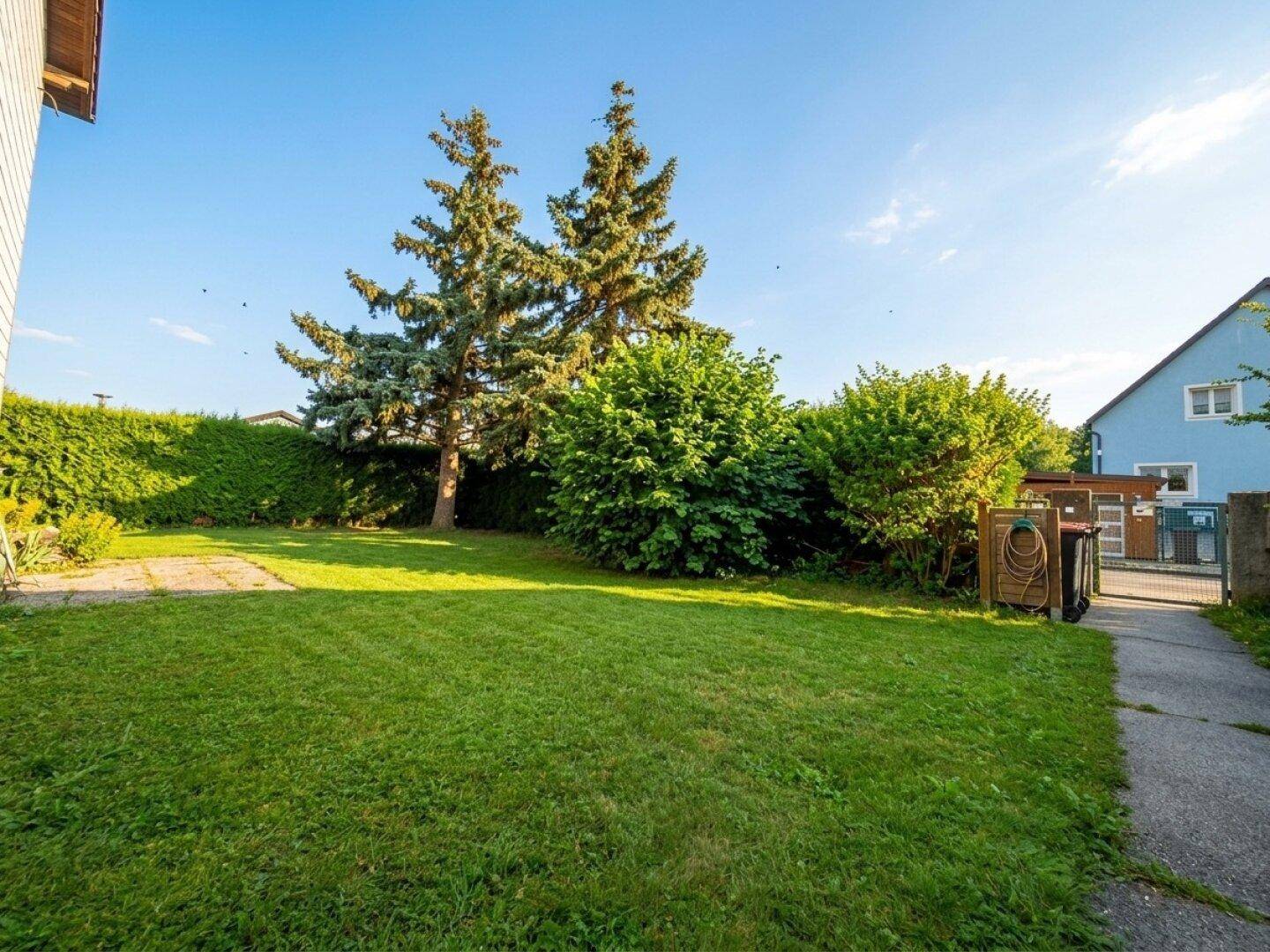 Großer, gepflegter Gartenbereich mit Rasenfläche, Bäumen und Hecke unter blauem Himmel.