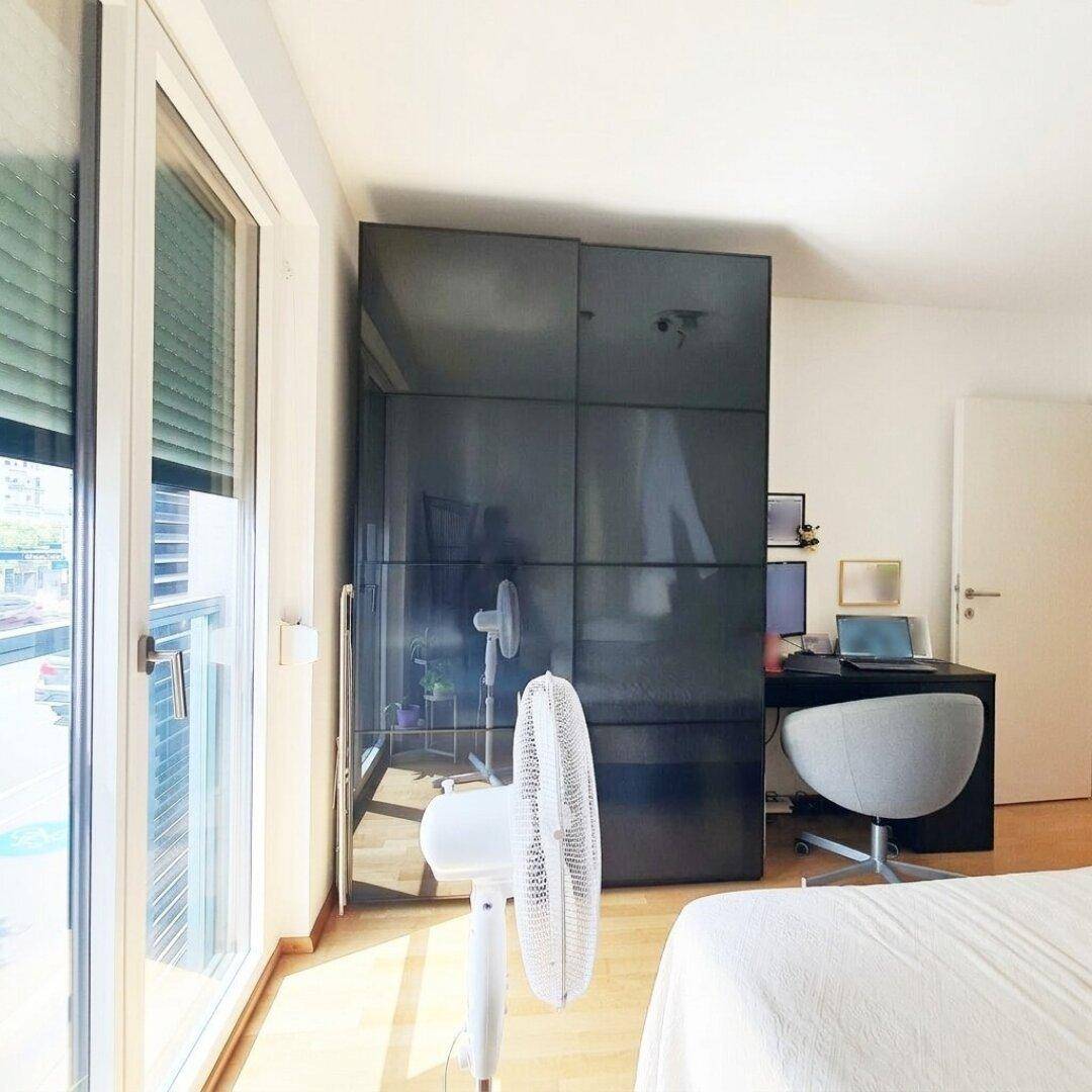 Helles Schlafzimmer mit großem Fenster, Schreibtisch und modernem Kleiderschrank.